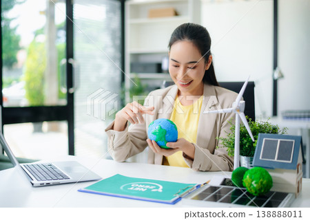 Asian woman working on laptop, holding green planet model with wind turbine and solar home nearby. Concept of clean energy 138888011