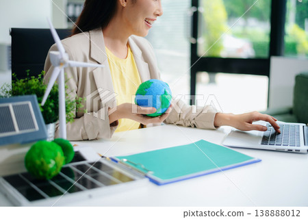 Asian woman working on laptop while holding green earth ball. Surrounded by wind turbine, solar panel, and eco-home models. Concept of clean energy and sustainability. 138888012