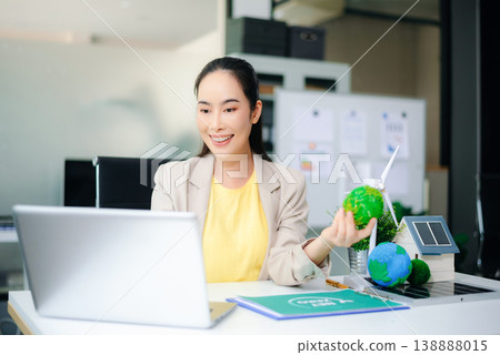 Asian woman working on laptop while holding green earth ball. Surrounded by wind turbine, solar panel, and eco-home models. Concept of clean energy and sustainability. Asian woman working on laptop while holding green earth ball. Surrounded by wind turbine, solar panel, and eco-home models. Concept of clean energy and sustainability. 138888015