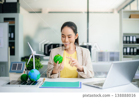 Asian woman working on laptop while holding green earth ball. Surrounded by wind turbine, solar panel, and eco-home models. Concept of clean energy and sustainability. 138888016