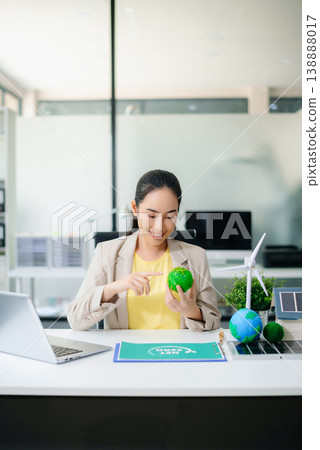 Confident Asian woman holding two symbolic Earth models at office desk with wind turbine and solar panel. Concept of sustainability, climate action, and clean energy business. 138888017