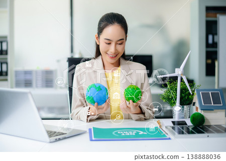 Confident Asian woman holding two symbolic Earth models at office desk with wind turbine and solar panel. Concept of sustainability, climate action, and clean energy business. 138888036