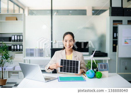 Asian woman holds solar panel in eco office with wind turbine, earth models, and net zero folder. Clean energy, renewable tech, and sustainability business concept. 138888040