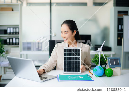 Asian woman holds solar panel in eco office with wind turbine, earth models, and net zero folder. Clean energy, renewable tech, and sustainability business concept. Asian woman holds solar panel in eco office with wind turbine, earth models, and net zero folder. Clean energy, renewable tech, and sustainability business concept. 138888041