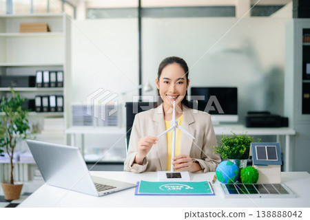 Young Asian woman working on laptop while holding wind turbine model. Surrounded by globe, solar panel, and green plants. Concept of sustainability and clean energy solutions. Young Asian woman working on laptop while holding wind turbine model. Surrounded by globe, solar panel, and green plants. Concept of sustainability and clean energy solutions. 138888042