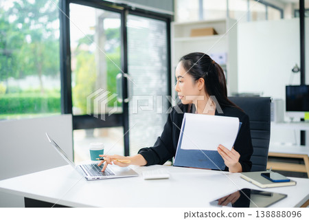 Smiling Asian businesswoman texting on phone at modern office desk with laptop, coffee, and documents Smiling Asian businesswoman texting on phone at modern office desk with laptop, coffee, and documents 138888096