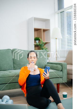 Fit woman in athleisure sits on yoga mat with tablet, headphones, and workout gear. Modern indoor wellness, healthy lifestyle, and digital fitness at home. 138888104