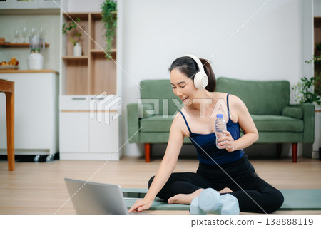 Asian woman practices yoga at home while watching an online tutorial on laptop. Indoor fitness, healthy lifestyle, stretching on mat in cozy modern living room. Asian woman practices yoga at home while watching an online tutorial on laptop. Indoor fitness, healthy lifestyle, stretching on mat in cozy modern living room. 138888119