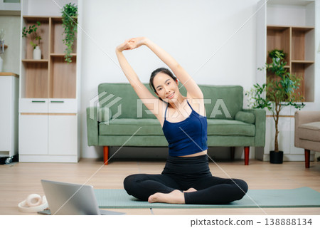 Young woman practices yoga and stretching with gym props on mat in modern home. Healthy lifestyle, wellness, motivation, balance, and indoor fitness activity. Young woman practices yoga and stretching with gym props on mat in modern home. Healthy lifestyle, wellness, motivation, balance, and indoor fitness activity. 138888134