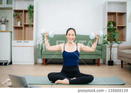 Young woman practices yoga and stretching with gym props on mat in modern home. Healthy lifestyle, wellness, motivation, balance, and indoor fitness activity. 138888139