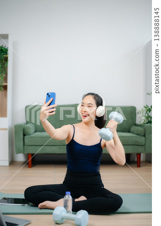 Young woman practices yoga and stretching with gym props on mat in modern home. Healthy lifestyle, wellness, motivation, balance, and indoor fitness activity. Young woman practices yoga and stretching with gym props on mat in modern home. Healthy lifestyle, wellness, motivation, balance, and indoor fitness activity. 138888145