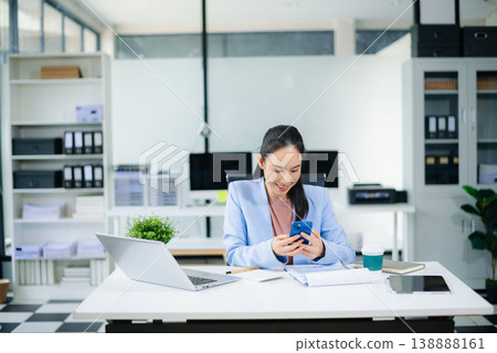 Asian businesswoman in blue blazer using tablet at modern office desk with laptop, coffee Asian businesswoman in blue blazer using tablet at modern office desk with laptop, coffee 138888161
