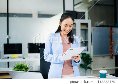 Asian businesswoman in blue blazer using tablet at modern office desk with laptop, coffee Asian businesswoman in blue blazer using tablet at modern office desk with laptop, coffee 138888170