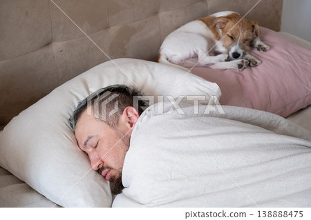Man sleeping in bed at home with his small dog. Caucasian guy resting on pillow next to pet puppy. Man sleeping in bed at home with his small dog. Caucasian guy resting on pillow next to pet puppy. 138888475