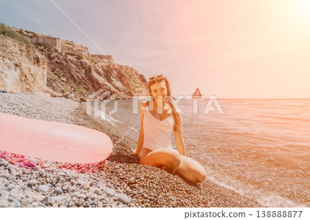 Beach girl swimsuit smiling on pebble shore by the sea with pink inflatable ring at sunset with cliffs in background 138888877