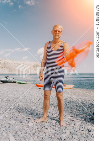 Man swimsuit beach stands on a pebble shore wearing a retro striped bodysuit while waving a vibrant orange fabric during summer vacation 138888878