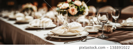 The wedding banquet table is laid out with plates and glassware. Floral arrangements add a touch of color. Guests will gather to celebrate in this beautiful space 138891022
