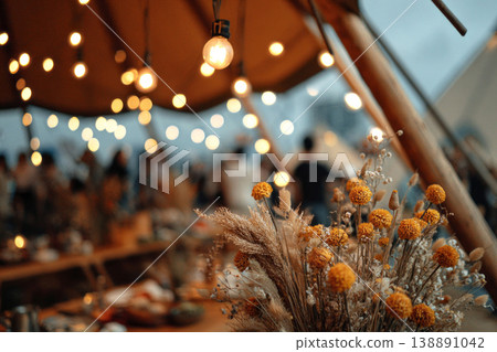 Guests gather under a large wedding tent filled with warm lights. The area is decorated with flowers and creates a festive ambiance for the outdoor celebration at dusk 138891042