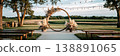 A wedding ceremony is set outdoors with a wooden arch adorned with wildflowers and pampas grass. Chairs are arranged in front of the arch. String lights hang in the background 138891065