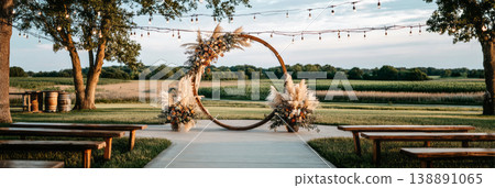 A wedding ceremony is set outdoors with a wooden arch adorned with wildflowers and pampas grass. Chairs are arranged in front of the arch. String lights hang in the background A wedding ceremony is set outdoors with a wooden arch adorned with wildflowers and pampas grass. Chairs are arranged in front of the arch. String lights hang in the background 138891065