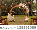 A wedding ceremony is set outdoors with a wooden arch adorned with wildflowers and pampas grass. Chairs are arranged in front of the arch. String lights hang in the background 138891069