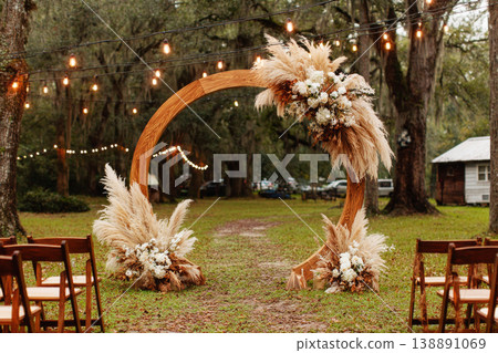 A wedding ceremony is set outdoors with a wooden arch adorned with wildflowers and pampas grass. Chairs are arranged in front of the arch. String lights hang in the background A wedding ceremony is set outdoors with a wooden arch adorned with wildflowers and pampas grass. Chairs are arranged in front of the arch. String lights hang in the background 138891069