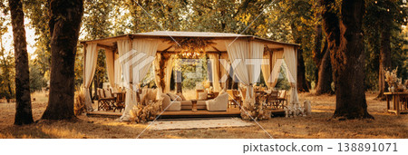 A wedding tent is set up in a grassy space with trees around. Inside, there are cushions and lights creating a cozy atmosphere for guests to relax and enjoy A wedding tent is set up in a grassy space with trees around. Inside, there are cushions and lights creating a cozy atmosphere for guests to relax and enjoy 138891071