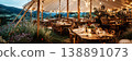 Guests are seated at wooden tables inside a wedding tent. Strings of lights hang from the top. The view shows water and city lights in the background during the evening 138891073