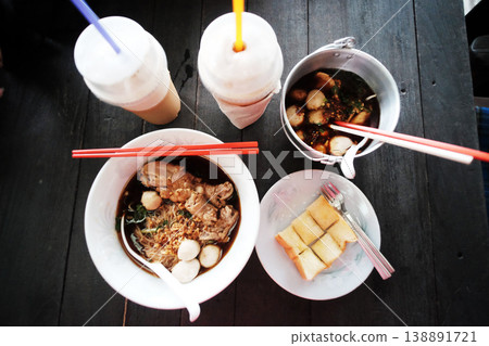 Top view of Thai boat noodles with chicken soup and pork meatballs in aluminum bowl with toast or bread with butter topping and blended latte coffee in plastic glass Top view of Thai boat noodles with chicken soup and pork meatballs in aluminum bowl with toast or bread with butter topping and blended latte coffee in plastic glass 138891721