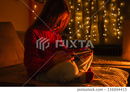 Girl in a red sweater playing a portable game device on a bed illuminated by warm string lights Girl in a red sweater playing a portable game device on a bed illuminated by warm string lights 138894415