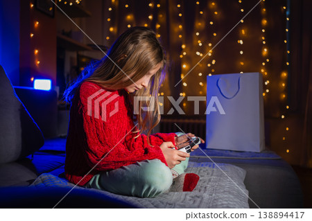 Girl sitting on a bed playing a game with blue lighting, warm garland, and a white paper bag visible Girl sitting on a bed playing a game with blue lighting, warm garland, and a white paper bag visible 138894417