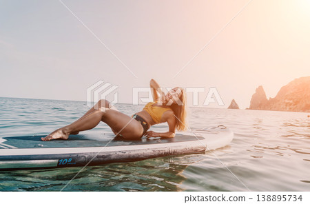 Woman Paddleboard Beach Relaxing: Young woman in a swimsuit relaxing on a paddleboard in the ocean near a beach. 138895734