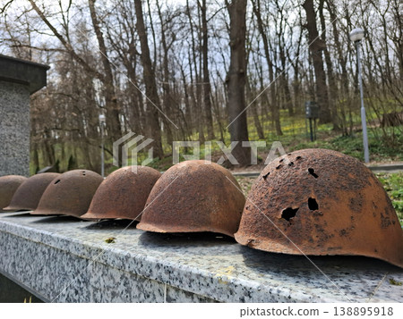 A row of old military helmets shows signs of wear with rust and bullet holes. These helmets sit on a stone ledge in a forest area surrounded by trees and greenery. 138895918