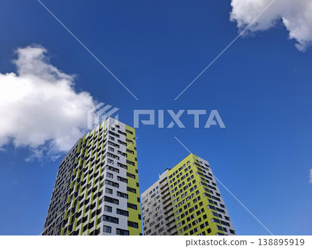 Bright modern apartment buildings stand tall under a clear blue sky. Fluffy clouds move gently above the colorful structures in an urban setting during daytime. 138895919