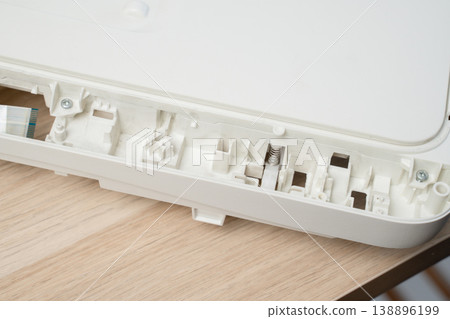 Open white printer and scanner casing interior plastic compartments, spring mechanism and ribbon cable on a wooden desk. Electronics repair, device maintenance, teardown and technical illustration. Open white printer and scanner casing interior plastic compartments, spring mechanism and ribbon cable on a wooden desk. Electronics repair, device maintenance, teardown and technical illustration. 138896199