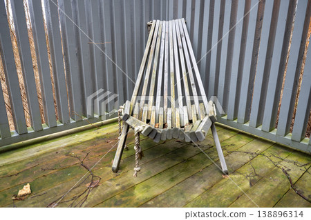Folded wooden deck chair on weathered terrace with gray fence and rustic outdoor texture 138896314