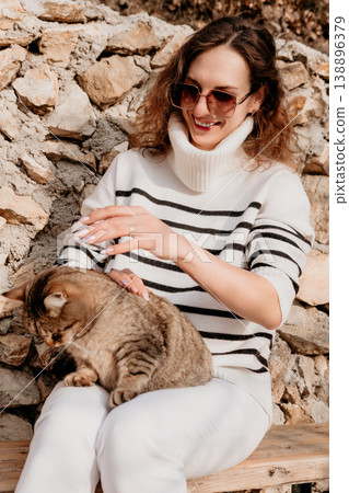Woman cat petting smiling lady in sunglasses sitting on a bench and stroking a tabby cat against a rustic stone wall Woman cat petting smiling lady in sunglasses sitting on a bench and stroking a tabby cat against a rustic stone wall 138896379