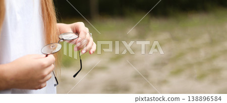 Close-up of a teenage girl holding round eyeglasses in her hands outdoors. Soft natural light, blurred green park background. Vision correction and ophthalmology concept for youth 138896584