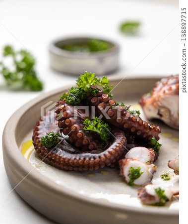 Boiled octopus tentacles and sliced pieces on ceramic plate close up. Olive oil and parsley dressing 138897715