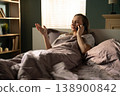 Woman talking on a mobile phone while lying comfortably in bed under a blanket, gesturing with her hand, communicating and receiving a call in her bedroom at home 138900842