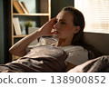Young woman lying in bed at dawn, pressing temples in pain and grimacing from a severe headache or migraine, morning light filtering into a quiet bedroom scene 138900852