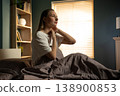 Woman sitting on bed in morning light touching her neck and shoulder, wincing from stiff neck and discomfort after waking up in her sunlit bedroom 138900853