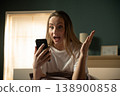 Woman in bedroom reacting with disbelief and astonishment while reading shocking news or an unexpected message on her smartphone, conveying emotional impact and unexpected information 138900858