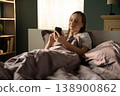 Woman lying awake in bed, comfortably checking her smartphone, connecting with the digital world on a sunny morning, reflecting modern daily routines and social media communication 138900862