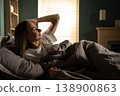 Woman waking up slowly in her bed, stretching her arm behind her head and enjoying the warm morning sunlight filtering through the window into her cozy bedroom 138900863