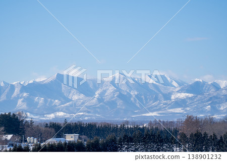 Hokkaido winter: Snow-capped mountain ranges visible beyond the town. A spectacular view representing the magnificent nature of Hokkaido. The snow-covered peaks shining in winter against a clear blue sky. 138901232