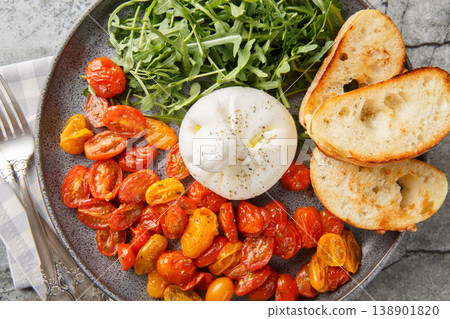 Delicious Italian burrata, baked cherry tomatoes and fresh arugula with olive oil and toasted ciabatta close-up in a plate. Horizontal top view 138901820
