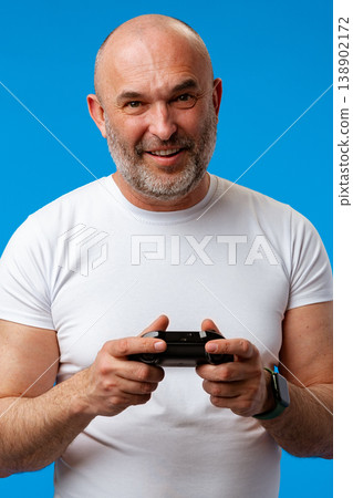 Older man enjoys video games with a controller in a bright blue background setting Older man enjoys video games with a controller in a bright blue background setting 138902172