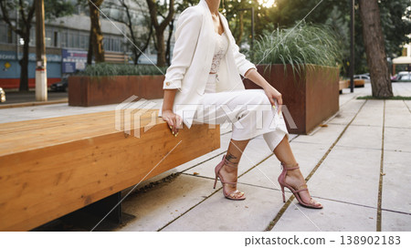 Woman sits on bench in city park wearing white outfit and pink heels during sunny day Woman sits on bench in city park wearing white outfit and pink heels during sunny day 138902183