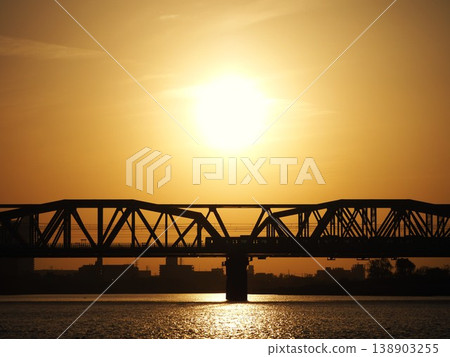 An orange sunset and the silhouette of a train crossing the Arakawa Railway Bridge. 138903255